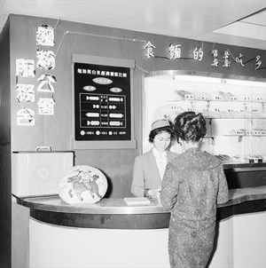 麵粉工會服務台前展示著掛著以一家七口平均計算之「麵粉與白米經濟價值比較」，一旁櫥窗展示著以麵粉製作之食品；服務台小姐正與婦女解說麵粉相關事宜。